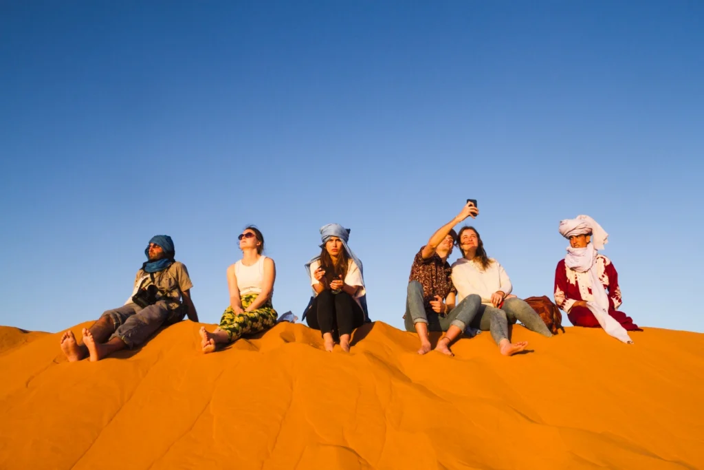 Group of travelers sitting on a sand dune at sunset, wearing comfortable and breathable desert safari outfits, highlighting casual examples of what to wear in Dubai desert safari.